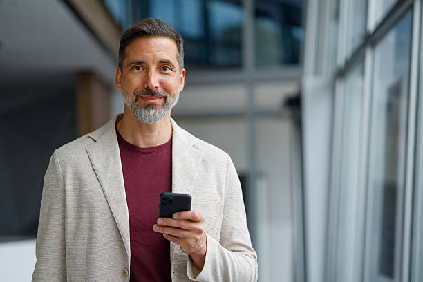 istock Confident mid adult businessman standing in a contemporary office, using mobile technology for remote work and connectivity 2240960876