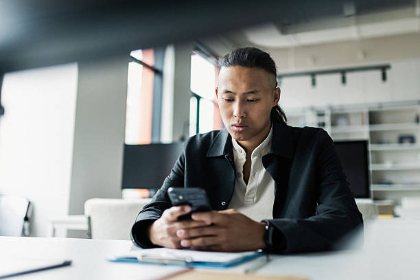 A focused business professional sits at a bright, minimalist office desk, intently using a smartphone. Papers and stationery create a productive, contemporary workspace atmosphere.