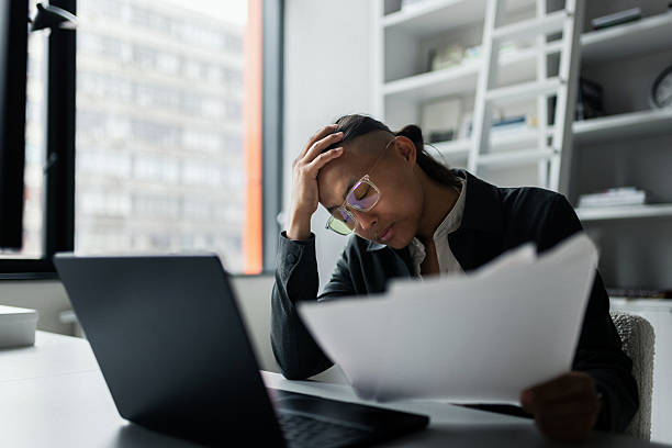 A focused professional sits at a desk, head in hand, reviewing documents beside a laptop in a bright office. The scene conveys stress, fatigue, and workload in a contemporary work environment.