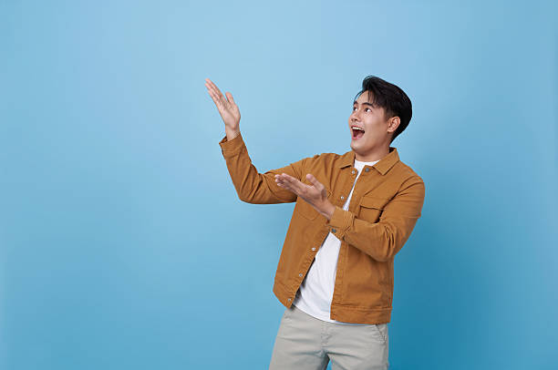 istock Asian man smiling and pointing finger sideways isolated on blue background representing presentation introduction creative idea and positive communication in modern lifestyle. 2240994153
