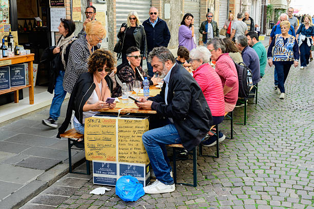 Outdoor café with diners enjoying their meals and drinks under a canopy in Barcelona, Spain. The scene features several tables with people seated, shaded by the café's awning, with a waiter attending to the patrons. The background includes adjacent buildings with traditional architectural details, and the sunlit street is lined with trees, adding to the lively atmosphere of the city.