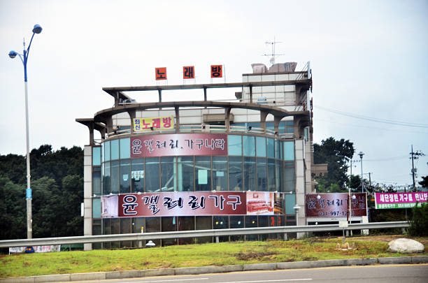 A commercial building in South Korea displays layered signage for Yoon Gallery Furniture and related businesses, including a karaoke bar and HVAC services. The glass façade and roadside placement reflect the spatial rhythm of suburban retail corridors, where furniture showrooms serve as both utility and visual anchor. Ganghwa Island, South Korea. August 23, 2013