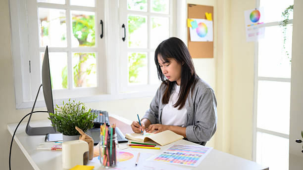istock Creative woman designing logo sketches on notebook with color samples and tools in bright modern workspace 2241024660