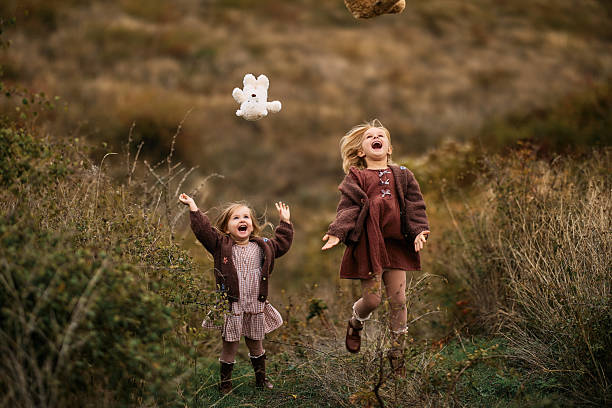 Two cheerful young girls laughing and jumping, throwing stuffed animals in a grassy outdoor environment