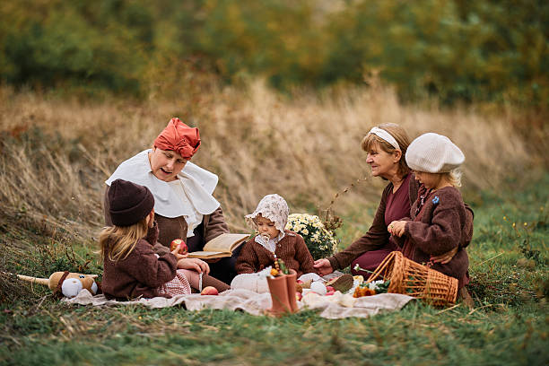 Family members in historical costumes enjoying an autumn picnic, older woman reading book to children