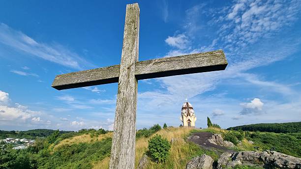 Wooden cross overlooking egg-shaped chapel on hill under bright sky, religious landscape in summer