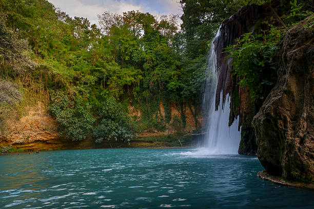 istock Tall natural waterfall plunging to blue river pool, surrounded by lush forest cliffs 2241049527