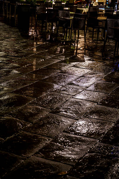 Night view of shiny, wet stone tiles reflecting lights near outdoor restaurant seating.