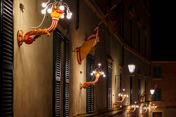 Bright, ornate wall lamps illuminate Italian windows and flags on a quiet night.