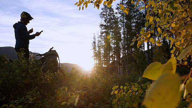 Backlit view of mature man with e-bike uses mobile phone in mountain meadow at sunrise, autumn leaves, Crowsnest Pass, Alberta