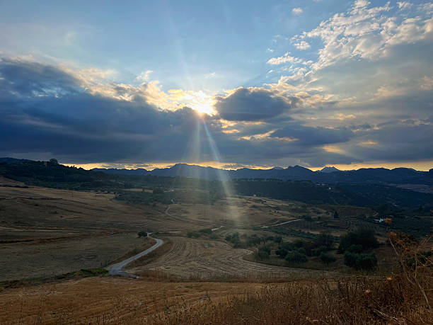 Hilly landscape with winding road under cloudy sky with sun shining through. Andalusia, Spain.