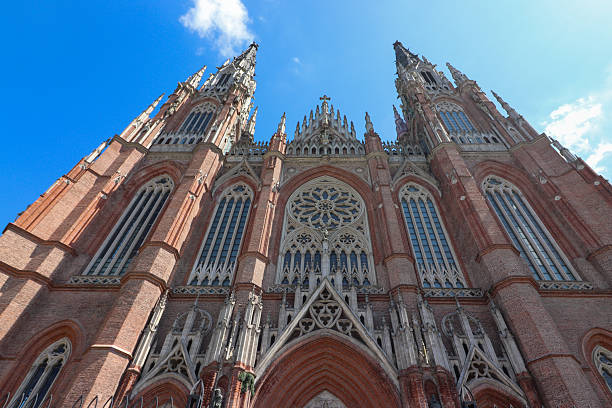 Dramatic vertical view of the La Plata Cathedral towers and spire against a bright blue sky with clouds. Focus on height, detail, and sun reflection.