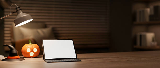 istock White screen tablet with halloween pumpkin lantern and witch hat under lamp on table in a dark room. 2241105747