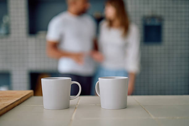 Coffee mugs on a tiled kitchen counter with a blurred couple standing in the background, having a morning conversation