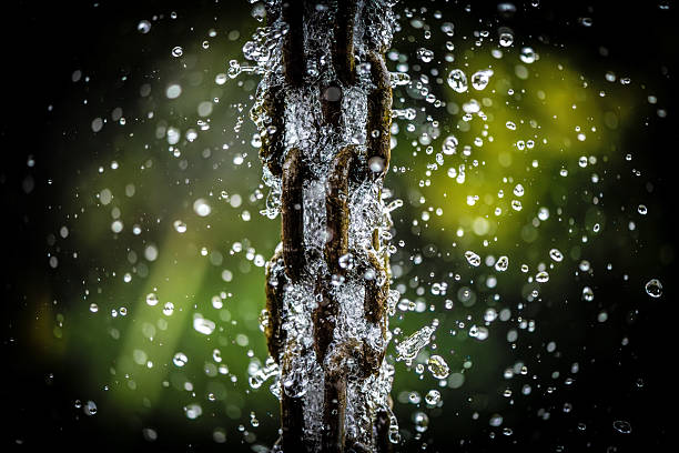 A close-up macro shot capturing water droplets splashing off a rusty metal chain. The photograph freezes the motion of the droplets with sharpness, revealing texture, reflections, and dynamic energy. The blurred green background enhances the contrast and gives depth to the composition.