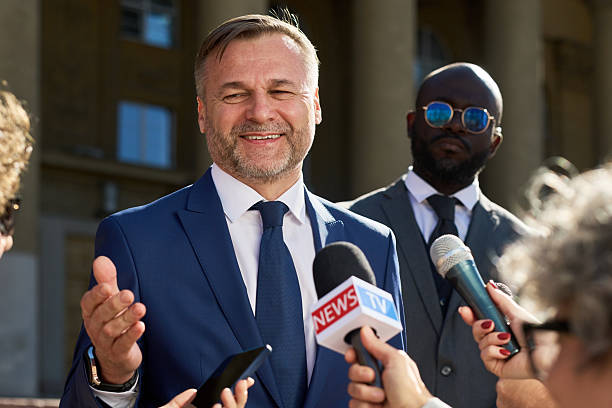 Caucasian middle aged man smiling and gesturing while speaking to journalists holding microphones outdoors, Black young man in suit standing behind observing press interaction