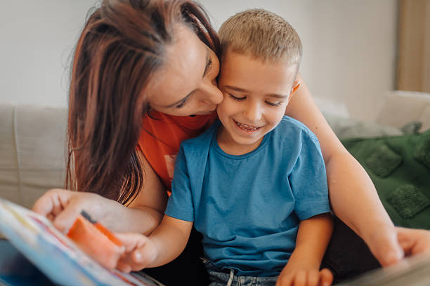 Happy mother kissing her son while reading a book together on the sofa at home, enjoying quality family time