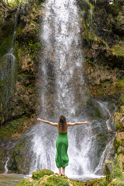 Charming young woman enjoying fresh air near waterfall at summer. She is wearing long green drees. Zlatibor mountain, Serbia