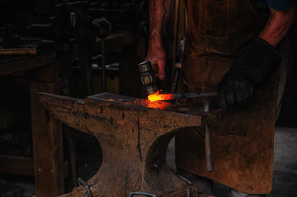 The blacksmith manually forging the molten metal on the anvil in smithy with spark fireworks
