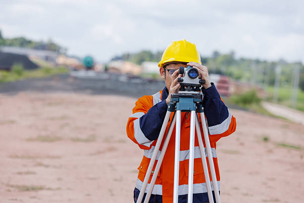 istock A survey engineer wearing an orange reflective jacket and yellow helmet operates a leveling instrument on a tripod at a construction site, symbolizing precision, measurement, and infrastructure development. 2241144138