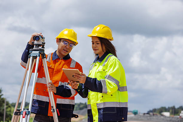 istock Two engineers wearing yellow safety helmets and reflective jackets discuss data on a digital tablet beside surveying equipment at a construction site. The scene reflects teamwork, precision, and technology. 2241144143