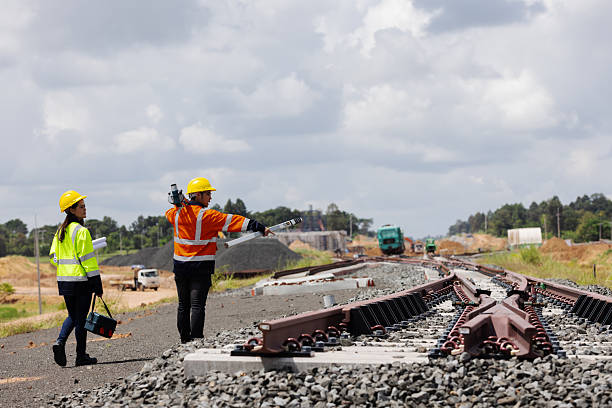 istock Two railway engineers wearing safety helmets and reflective vests inspect new rail tracks using surveying equipment. The image shows construction progress, precision, and teamwork under cloudy skies. 2241144148