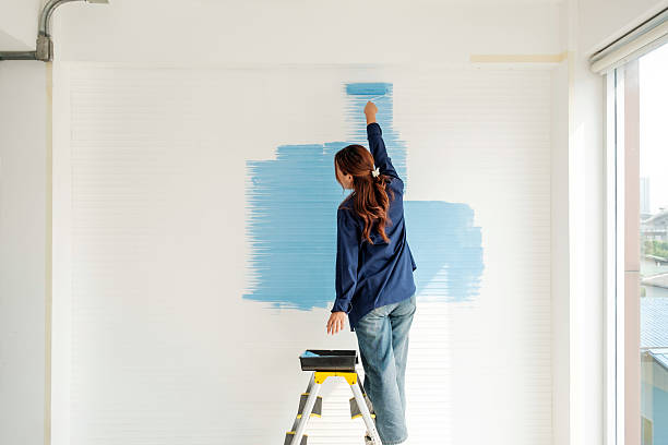 istock Woman on step ladder paints white interior wall blue in bright home. Scene captures DIY painting, renovation, remodeling, redecorating, and apartment makeover during home improvement project. 2241145771