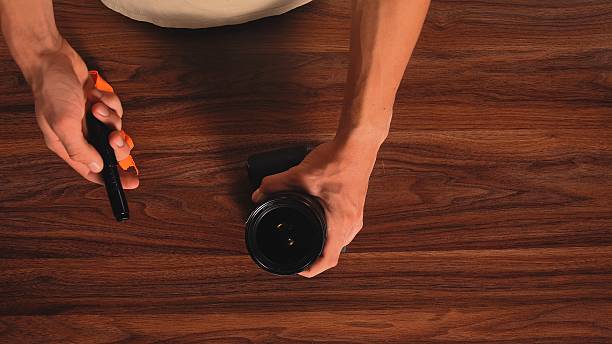 Top shot at hands cleaning a camera lens with a pen brush, placed on a wooden surface. The pen has an orange cloth for effective lens cleaning.