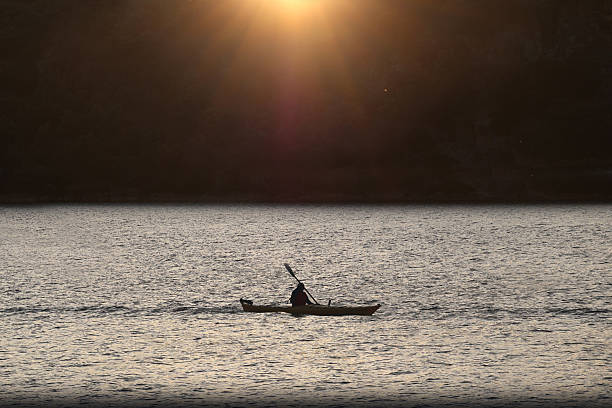 istock A man kayaking on a calm lake, with a backdrop of lush green trees and a distant mountain range. 2241153148