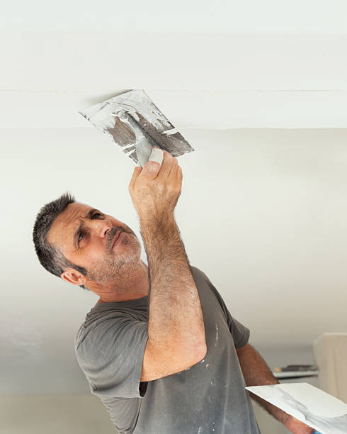 istock Man in grey work t-shirt uses trowel and hawk to plaster ceiling, construction or renovation of house 2241153500