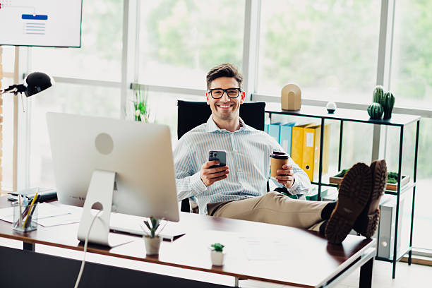 istock Confident young professional in a modern office relaxing at a desk with computer and smartphone while drinking coffee 2241153748
