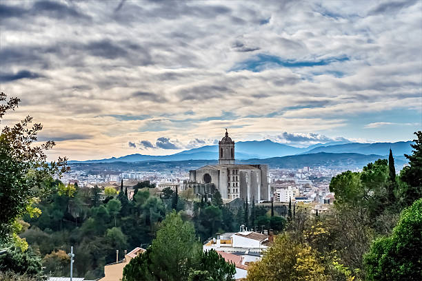 Panoramic view of Girona Cathedral with dramatic cloudy sky and mountains in the background, Spain. Historic city of Girona surrounded by lush greenery and cypress trees in Catalonia