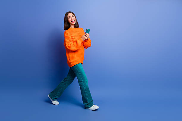 istock Young woman in bright orange sweater smiling and using smartphone while standing against vibrant blue background 2241155368