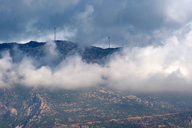 Wind turbines stand on a mountain ridge partially covered by mist. Trees and dense vegetation fill the foreground, while clouds drift across the slopes, blending nature with renewable energy in a tranquil scene.