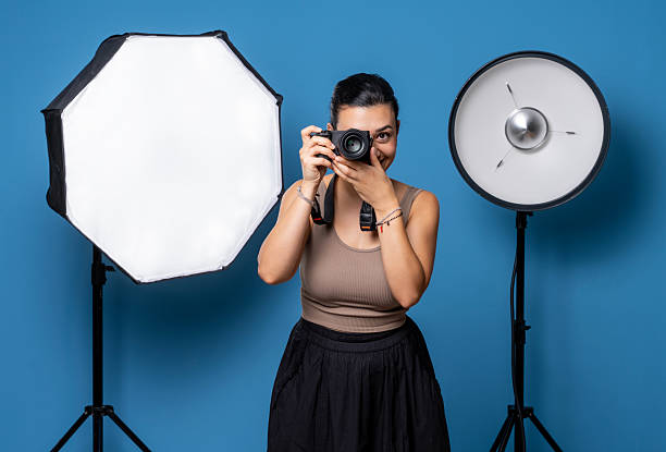 A woman photographer holding a camera and taking a picture in a studio, standing between two professional softbox lights.