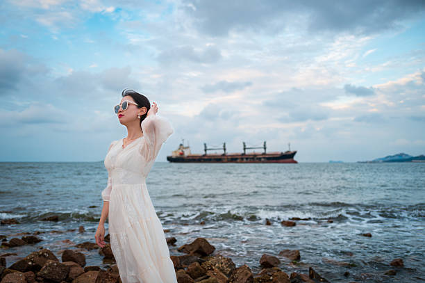 A woman in a white long dress walking towards the seaside, with her back to the camera. In the distance, a large cargo ship is quietly docked on the water, as gentle waves lap against the shore, creating a serene and poetic atmosphere
