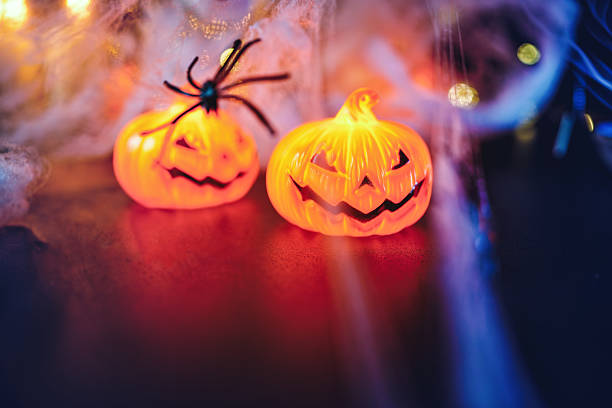 Two glowing pumpkin lights with cobwebs and a black spider on a dark surface. The bright orange glow and blurred background evoke a sense of playful eeriness and seasonal celebration. Copy space for text - Halloween, Thanksgiving