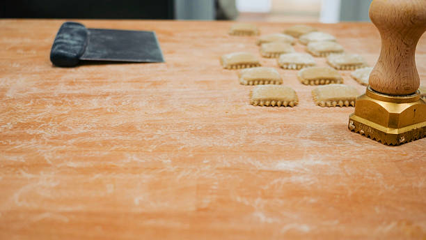 Artisanal pasta making process close up showing rows of fresh ravioli being cut and stamped on a wooden table dusted with flour alongside a metal spatula and a wooden rolling pin