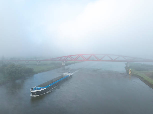 Ship passing the Hanzeboog train bridge over the river IJssel with during a foggy autumn morning and a train of the NS passing over the red bridge near the city of Zwolle in Overijssel, The Netherlands. Aerial drone point of view.