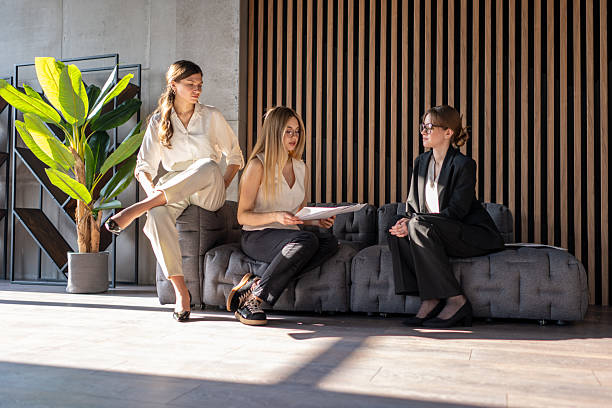 Three businesswomen are sitting on a modern office sofa, discussing important partnership details and collaborating on paperwork, representing teamwork and professional communication