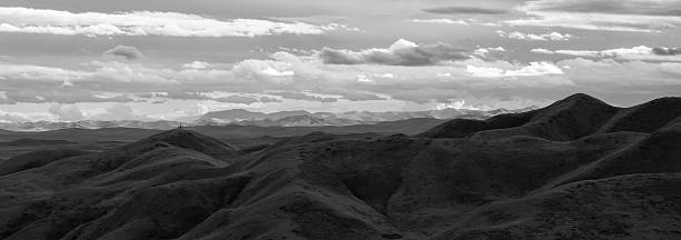 Monochrome panoramic photograph of undulating hills stretching into the distance beneath a dynamic sky filled with layered clouds. The soft contours of the terrain contrast beautifully with the textured clouds, evoking a sense of solitude and timelessness. Ideal for illustrating themes of nature, wilderness, serenity, geography, exploration, or minimalism.
