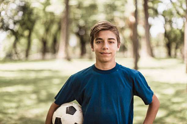 Description: A young soccer player in a blue shirt poses confidently with a soccer ball in a park setting, showcasing enthusiasm for the sport on a sunny day.