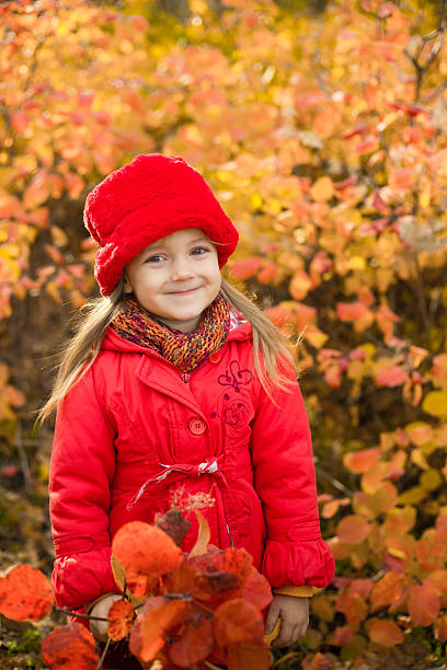 A mother and daughter wearing matching red hats enjoy a peaceful walk through a colorful autumn forest, holding hands and gathering leaves among golden and orange shrubs, capturing warmth, connection, and cozy fall vibes.