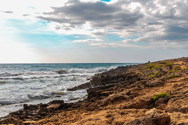 Golden coastal rock formations shaped by erosion, with ocean waves hitting the rugged shoreline under a partly cloudy sky. High quality photo