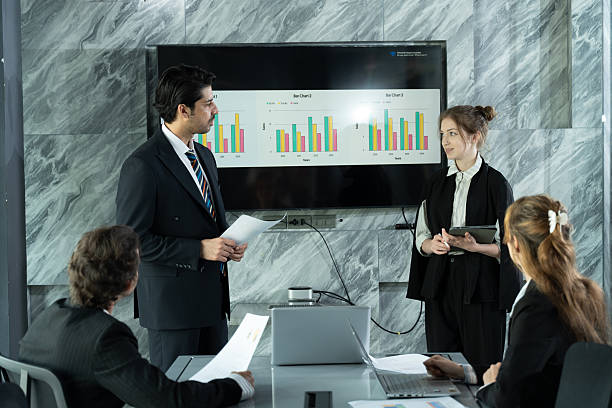 istock Caucasian businessman standing and explaining business chart during office meeting, multiracial colleagues sitting around table, female executive listening while typing on laptop inside conference room 2241196686