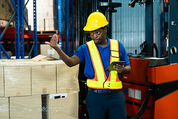 istock Adult African male warehouse worker explaining logistics tasks using digital tablet, standing in industrial storage area, wearing hard hat and vest, focusing on inventory coordination. 2241203753