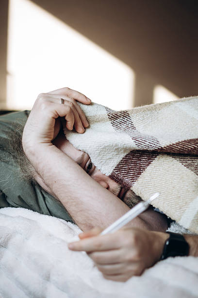 istock Man lying in bed at home, covered with a blanket, looking at a thermometer. 2241228920