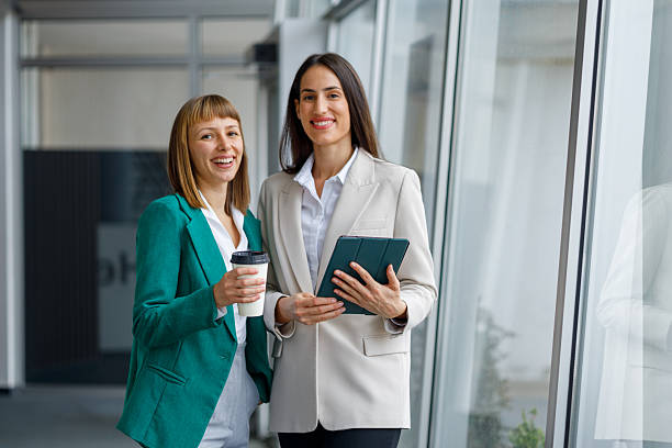 istock Professional businesswomen smiling during coffee break, holding tablet and discussing work in modern office corridor 2241235228