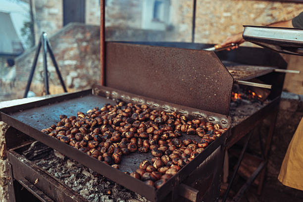 Close-up of a craftsman roasting chestnuts on a large iron plate over glowing embers. The rustic setting and warm tones capture the essence of autumn traditions and the timeless scale of artisanal food preparation.