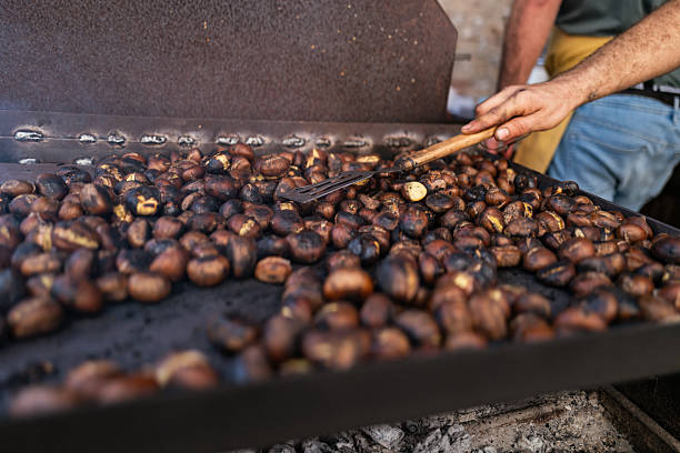 Close-up of a craftsman roasting chestnuts on a large iron plate over glowing embers. The rustic setting and warm tones capture the essence of autumn traditions and the timeless scale of artisanal food preparation.